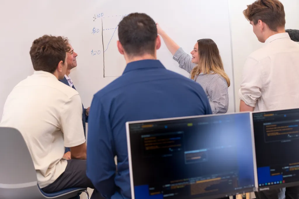 A student explains a rising stock price chart on a whiteboard to classmates, with financial data displayed on computer monitors in the foreground.