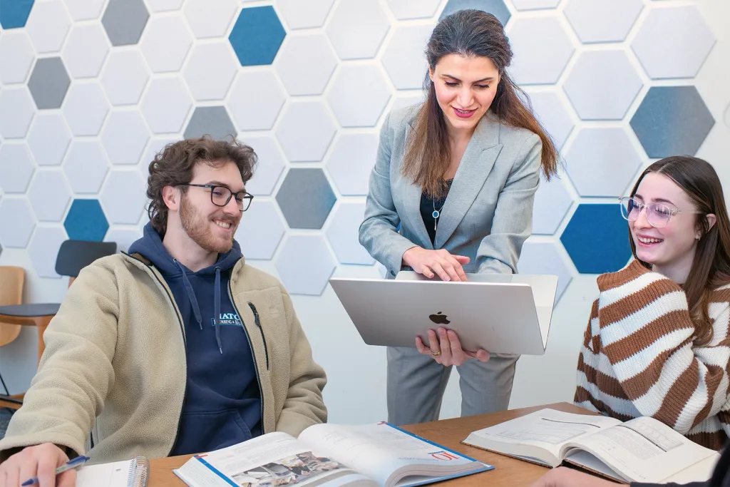 A professor holds a laptop while advising two smiling students seated at a table with open textbooks, in a commons area with a hexagon-patterned wall.