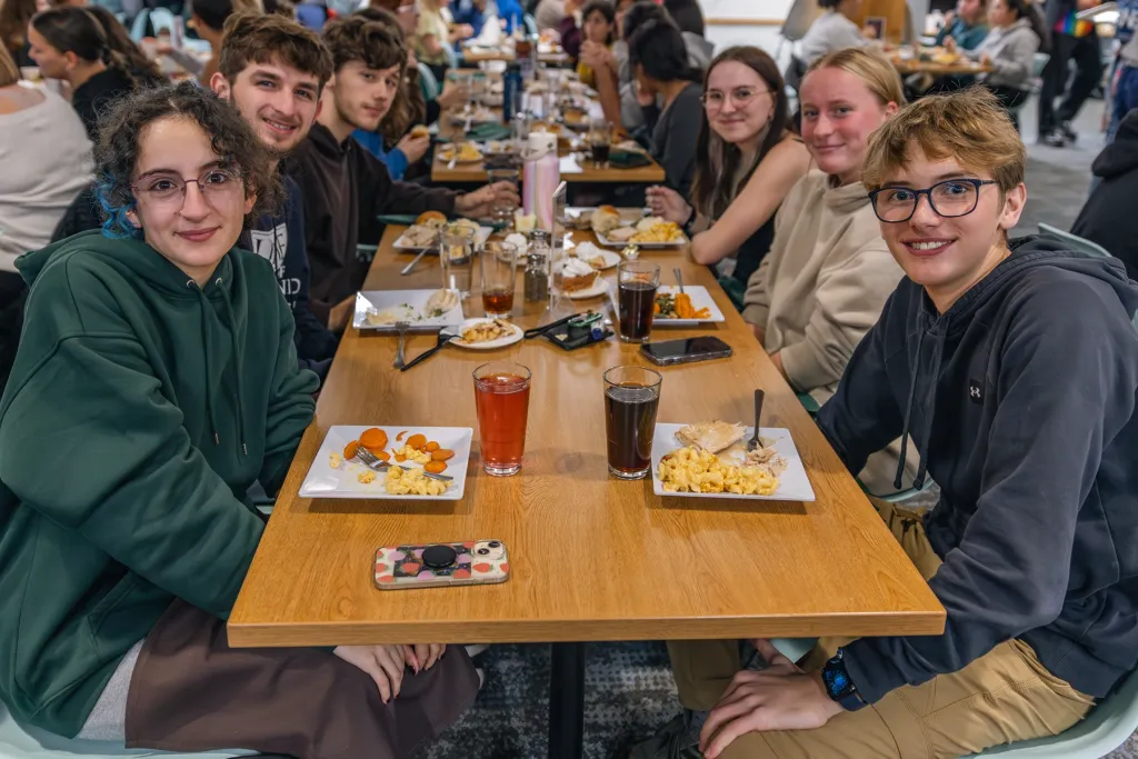 A group of students smiles at the camera while sharing a meal at a long table in the Commons.