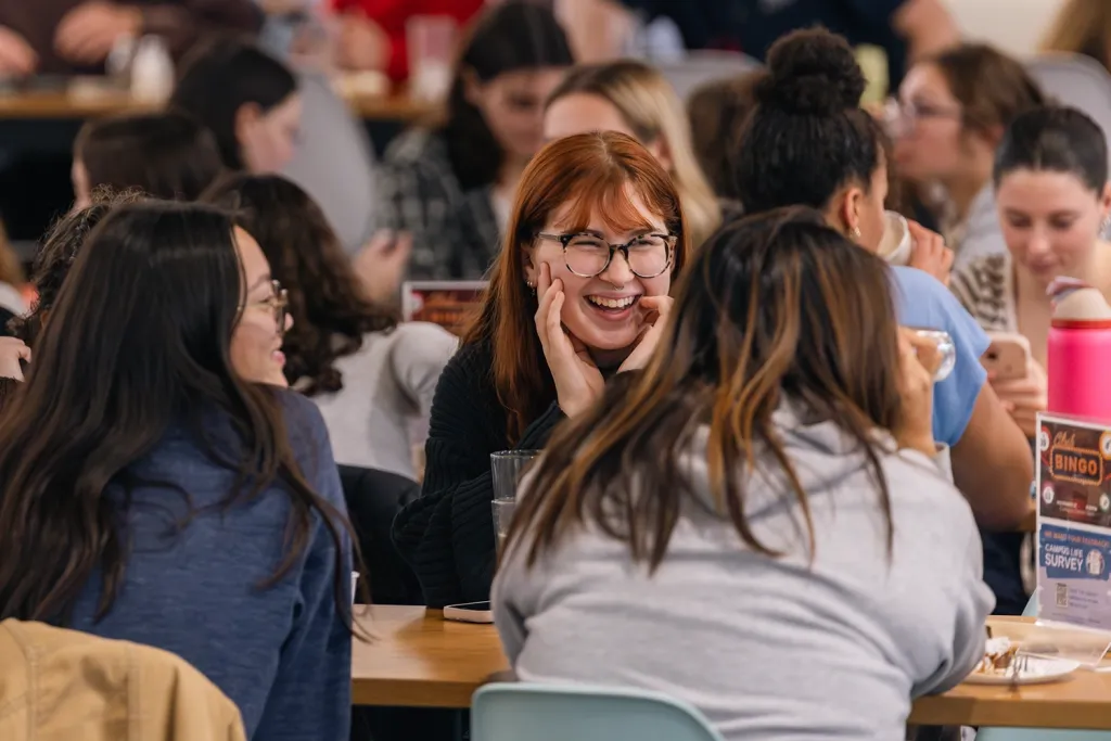 A student with red hair and glasses laughs with friends at a busy dining table in the Commons.