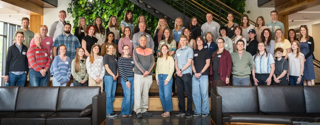 Group photo of the 2026 U N E Sustainability Fellows wearing name badges, gathered on a wooden staircase in front of a living plant wall.