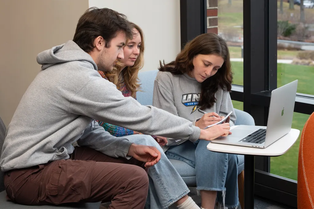 A young man in a gray hoodie points to the screen while a woman writes in a notebook; a second woman wearing a U N E Nor'easters sweatshirt looks on. Large windows behind them show a grassy campus lawn.