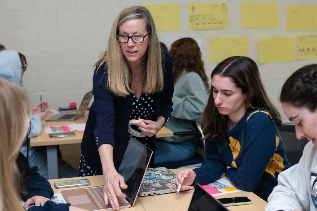 A professor leans over a student's desk to help with a laptop during a hands-on class activity. Students at nearby tables work at laptops covered in stickers, with yellow sticky notes and a workshop diagram posted on the wall behind them.