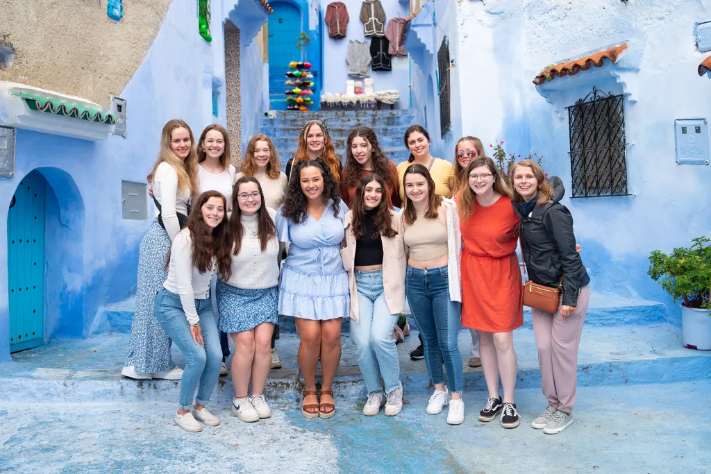 A group of U N E study abroad students pose together on blue-painted stairs in Chefchaouen, Morocco. The building's vivid blue walls and archways surround them, with woven baskets for sale in the background.