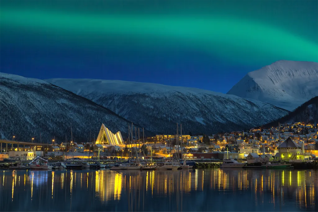 The northern lights glow green over Tromsø, Norway, illuminating the Arctic Cathedral and harbor at night.
