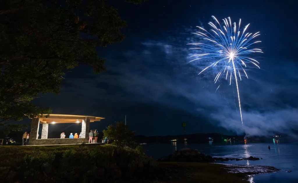 A firework bursts over the water at Jordan Point on U N E's campus as onlookers watch from a lit pavilion.