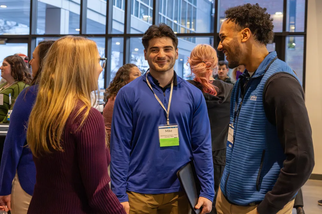 A U N E student wearing a name badge smiles while networking with an alum at an indoor mixer event.