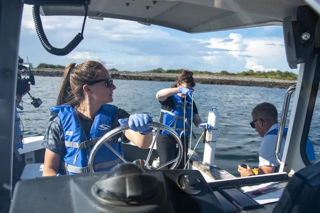 A U N E graduate marine science student steers a research boat while classmates collect water samples nearby.