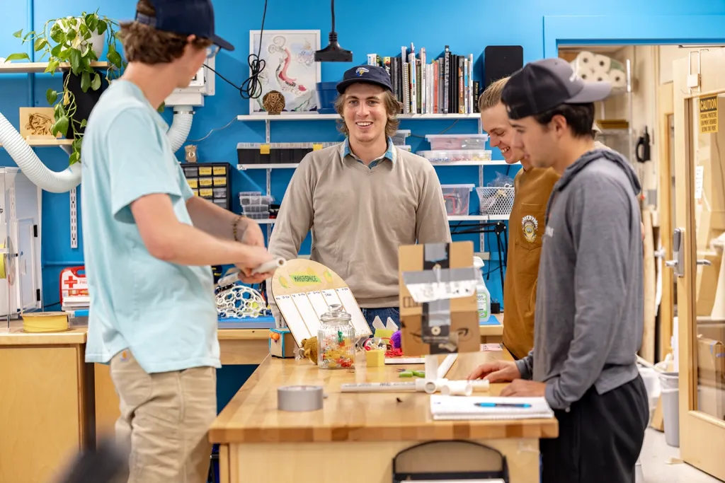 A group of U N E students gather around a workbench with craft materials and tools in the campus makerspace.