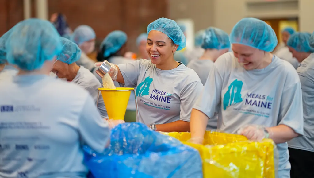 Volunteers in hairnets smile while packing bags of food at U N E's 2025 Meals for Maine event addressing food insecurity.