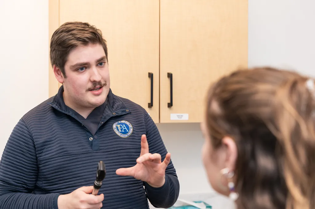 A U N E Physician Assistant student holding an otoscope explains something to a patient in an exam room.