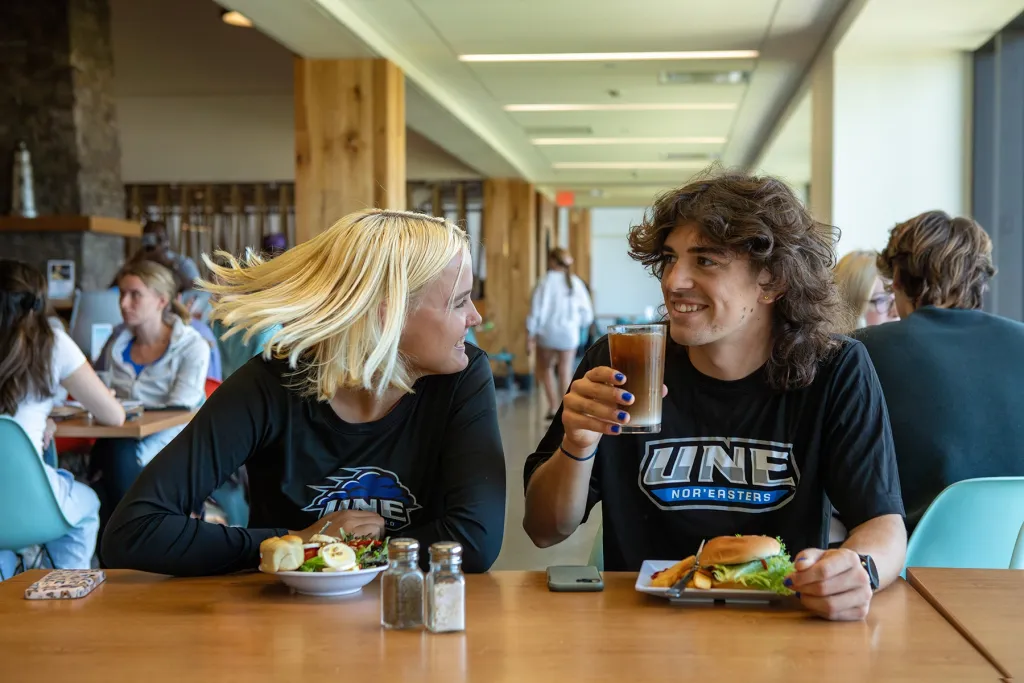 Two U N E student-athletes in Nor'easters shirts chat over lunch in the Commons dining hall.