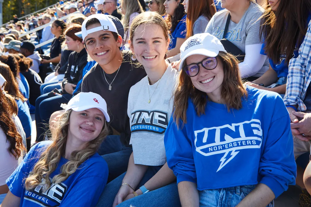 Four U N E students in U N E gear smile for the camera from the stands at a Homecoming football game.