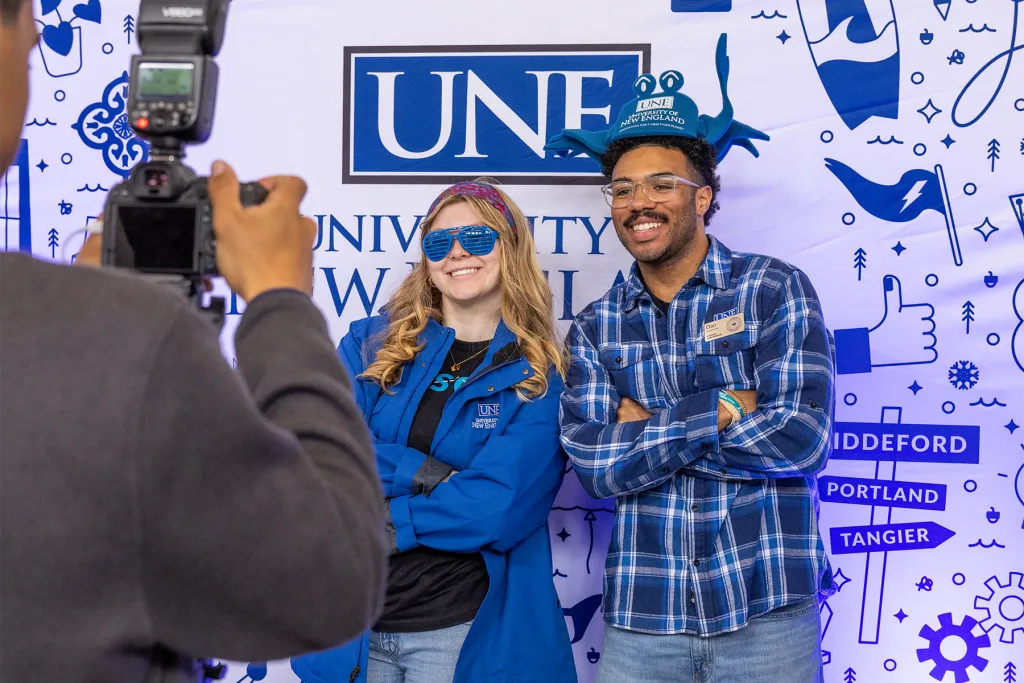 Two U N E students wearing silly props pose for a photo in front of a branded U N E step-and-repeat backdrop.