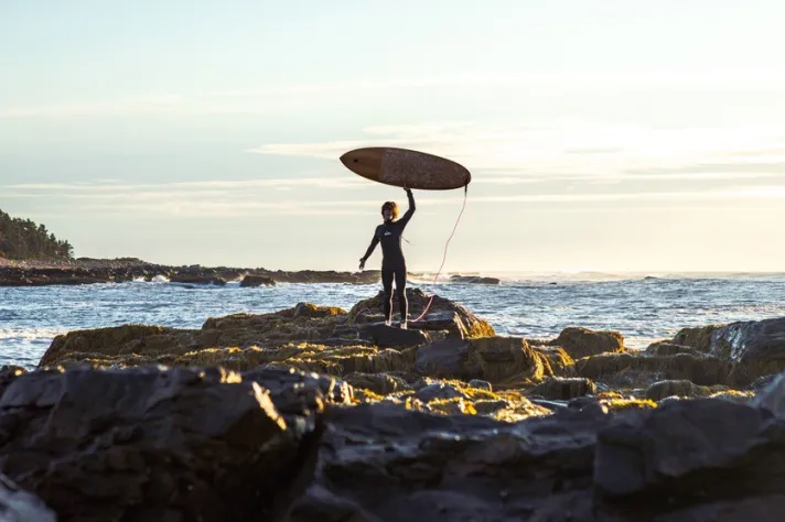 A student in a wetsuit holds up a surfboard from the top of a rocky coastline on the ocean
