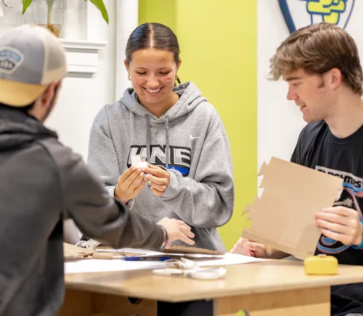 Student in UNE hoodie examining small white device while collaborating with classmates at table.