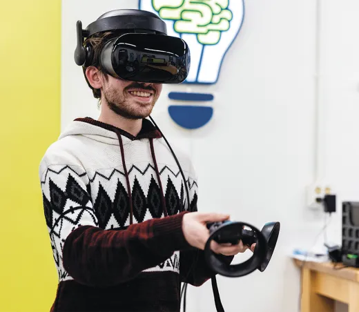 A student wearing a virtual reality headset and geometric-patterned hoodie grins while holding VR controllers in a makerspace.