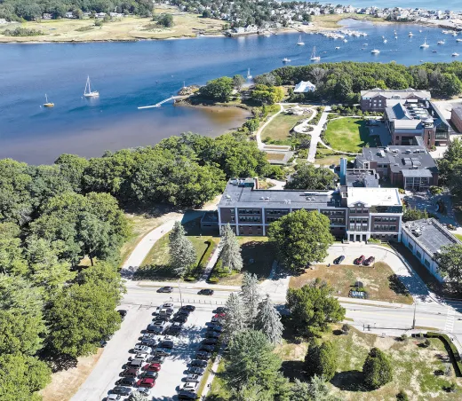 Aerial view of the University of New England's Biddeford Campus showing waterfront academic buildings, sailboats in the harbor, and the site where The Wayside once stood.
