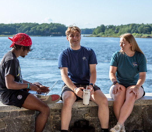 Three U N E students sit by the water on the Biddeford Campus
