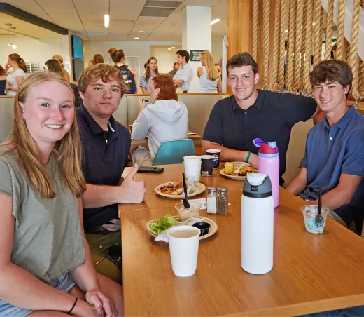 Five students smile together at a table in the campus dining commons, with food and drinks in front of them.