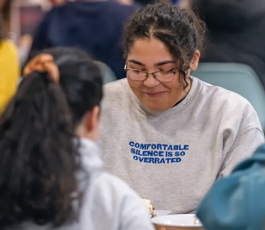 A student wearing glasses smiles while chatting with a friend across the table in the dining commons.