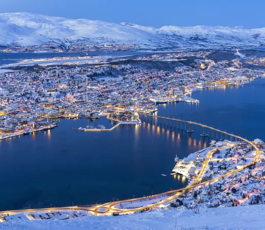 A dusk aerial view of Tromsø, Norway, showing the illuminated city, bridge, and fjord surrounded by snow-covered mountains.