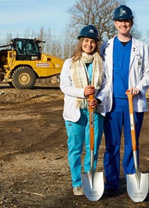 Two UNE COM students pose with shovels, the construction site in the background