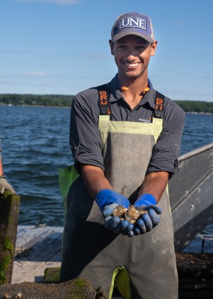 Michael Scannell in waders and a U N E baseball cap holds oysters in his hands while out on a boat
