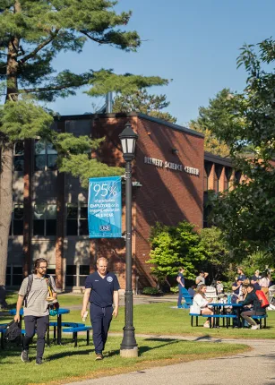 Two students walk past a green tree while other students sit at a picnic table outside of a brick building