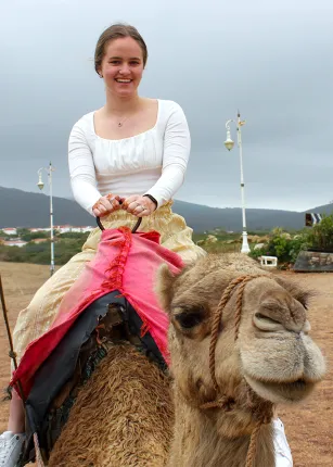 A student sitting on top of a camel with desert and mountains behind them