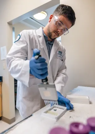 A student in safety gear and a white coat uses a multi-channel pipette to place liquid in a tray