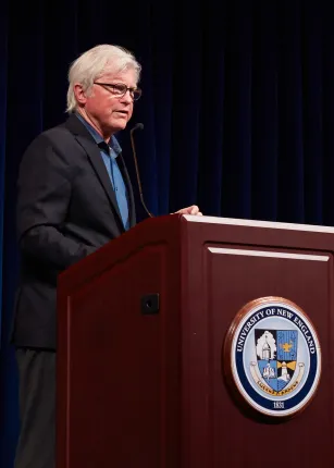 David Evans Shaw delivers a keynote address at a U N E podium as CEO of Blackpoint Group, with a presentation slide displayed on screen and attendees seated on stage behind him