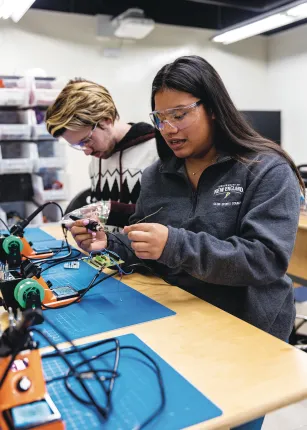A student wearing a University of New England quarter-zip and safety glasses works with soldering equipment and electronic components on a blue work mat.