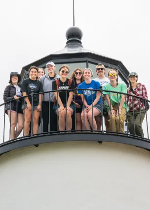 Group of students standing on the exterior gallery of a lighthouse lantern room, smiling at the camera against an overcast sky.
