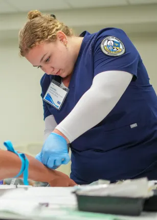 Nursing student wearing UNE scrubs and blue gloves practices clinical skills with medical equipment during hands-on training.