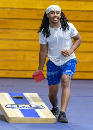 A student plays cornhole in the gymnasium with yellow bleachers in the background.