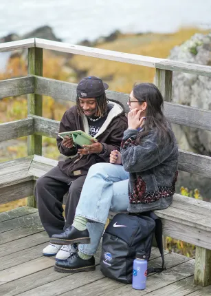 Two UNE students sit together on a wooden overlook, reviewing notes on a tablet with rocky coastal scenery in the background.