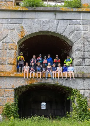 A large group of U N E students pose together inside a granite archway at Fort Gorges, sitting and standing on the historic fort's stone ledge.