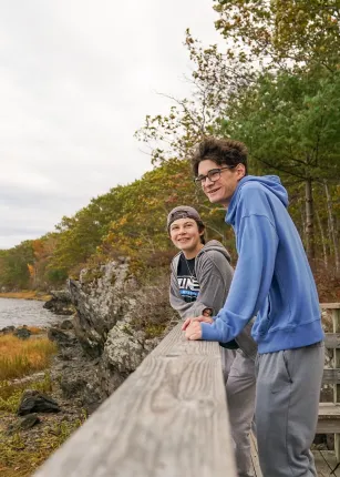 Two U N E students lean on a railing overlooking the rocky shoreline near Jordan's Point, surrounded by fall foliage.