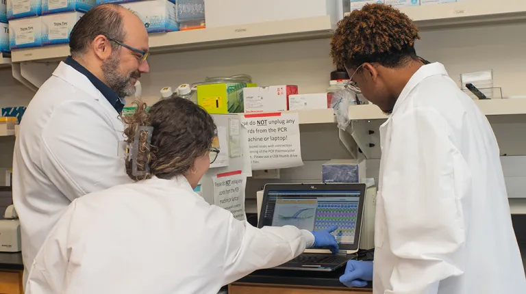 Two U N E students and a professor review research data on a computer