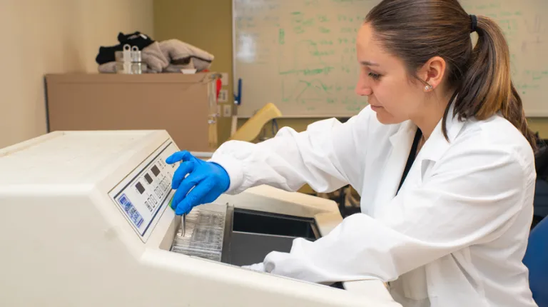 A student in a white lab coat works on a lab machine