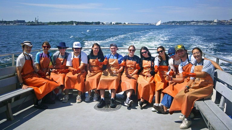 A group of students in fishing apparel sit on a boat in Casco Bay