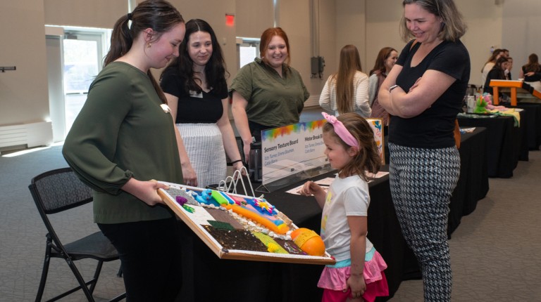 A group of students share a sensory texture board with a young girl at an Autism Expo