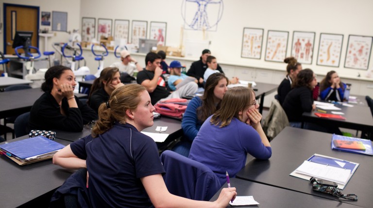 A group of athletic training students in a classroom with skeletomuscular portraits hanging on the wall