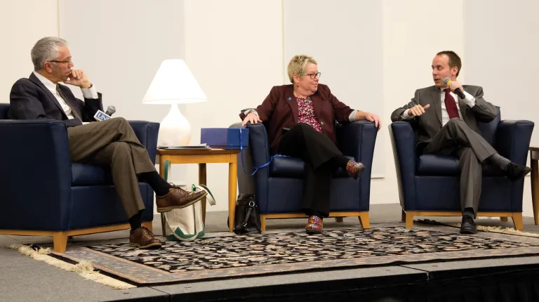 Three speakers sit in leather chairs and talking on the stage at a President's Forum