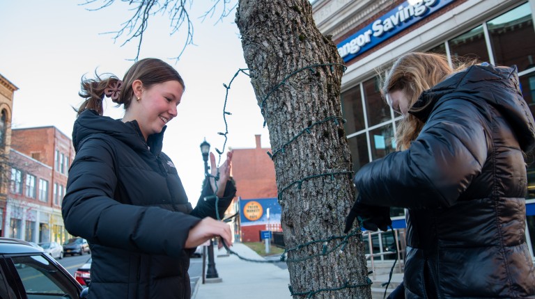 Two female students wrap holiday lights around the trunk of a tree in downtown Biddeford