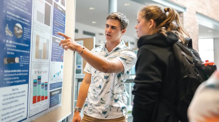 A student points to his research poster and discusses his findings with another student
