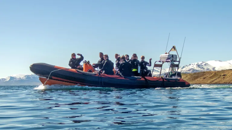 U N E students in wetsuits sit on a small boat with snow-covered mountains in the background