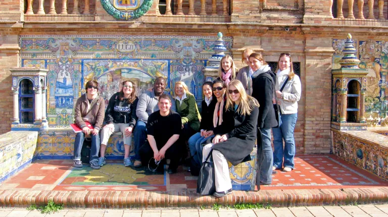 A group of students pose together in front of a building made with colorful tiles and brick