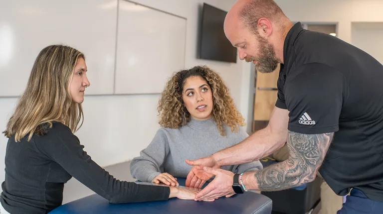 A professor helps an O T student measure the wrist of another student at an examination table